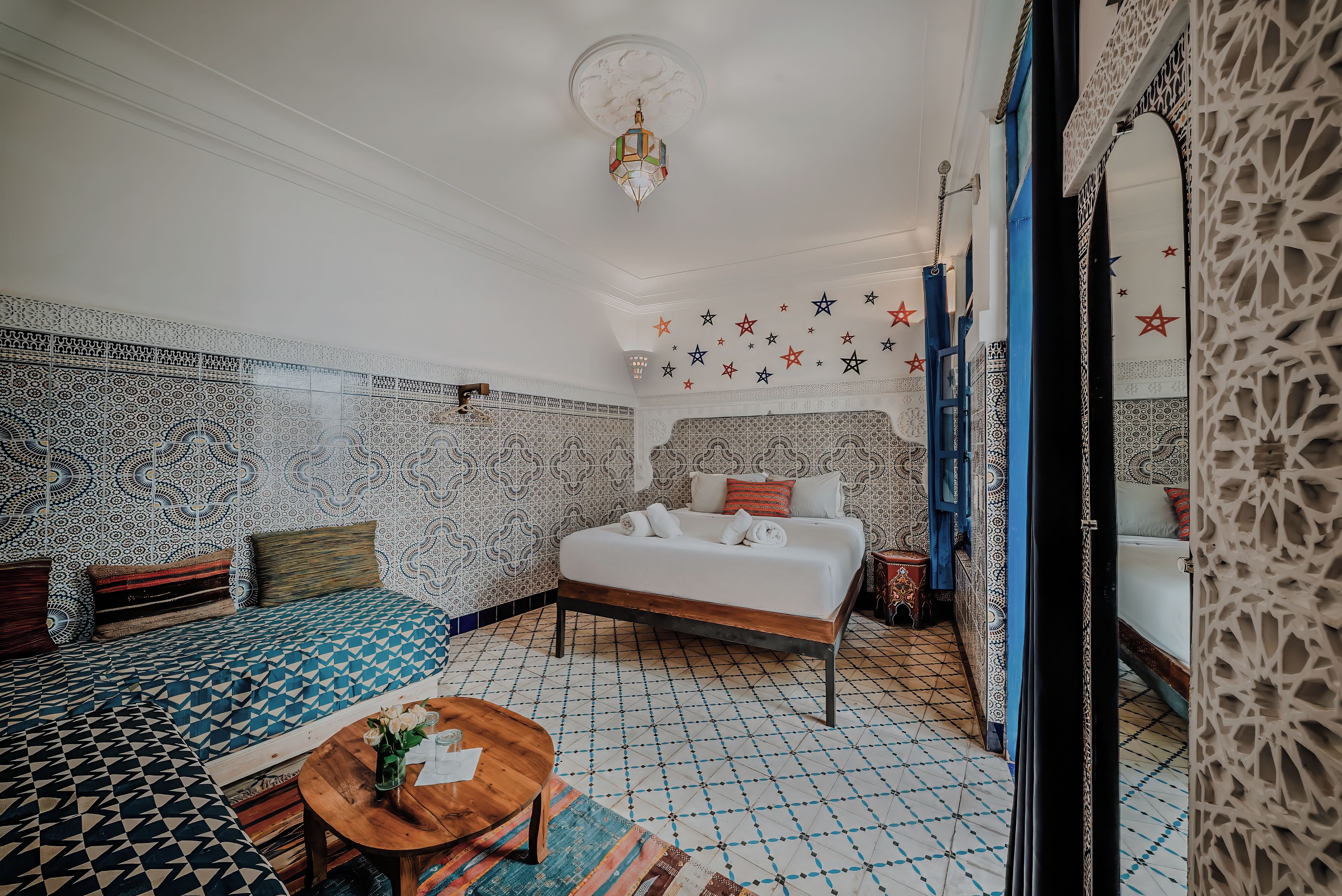 Una habitación con cama, sofá, una mesita con un florero, paredes con diseño y una lámpara decorativa en el techo.