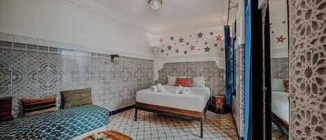 A room with a bed, a sofa, a small table with a vase, patterned walls, and a decorative ceiling light.