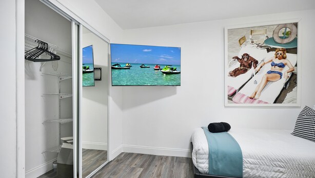 A bedroom with a bed, a closet with hanging clothes, and a wall-mounted TV displaying a beach scene. - Good Vibes Retreat Decor BBQ Patio heart Wynwood (Miami)