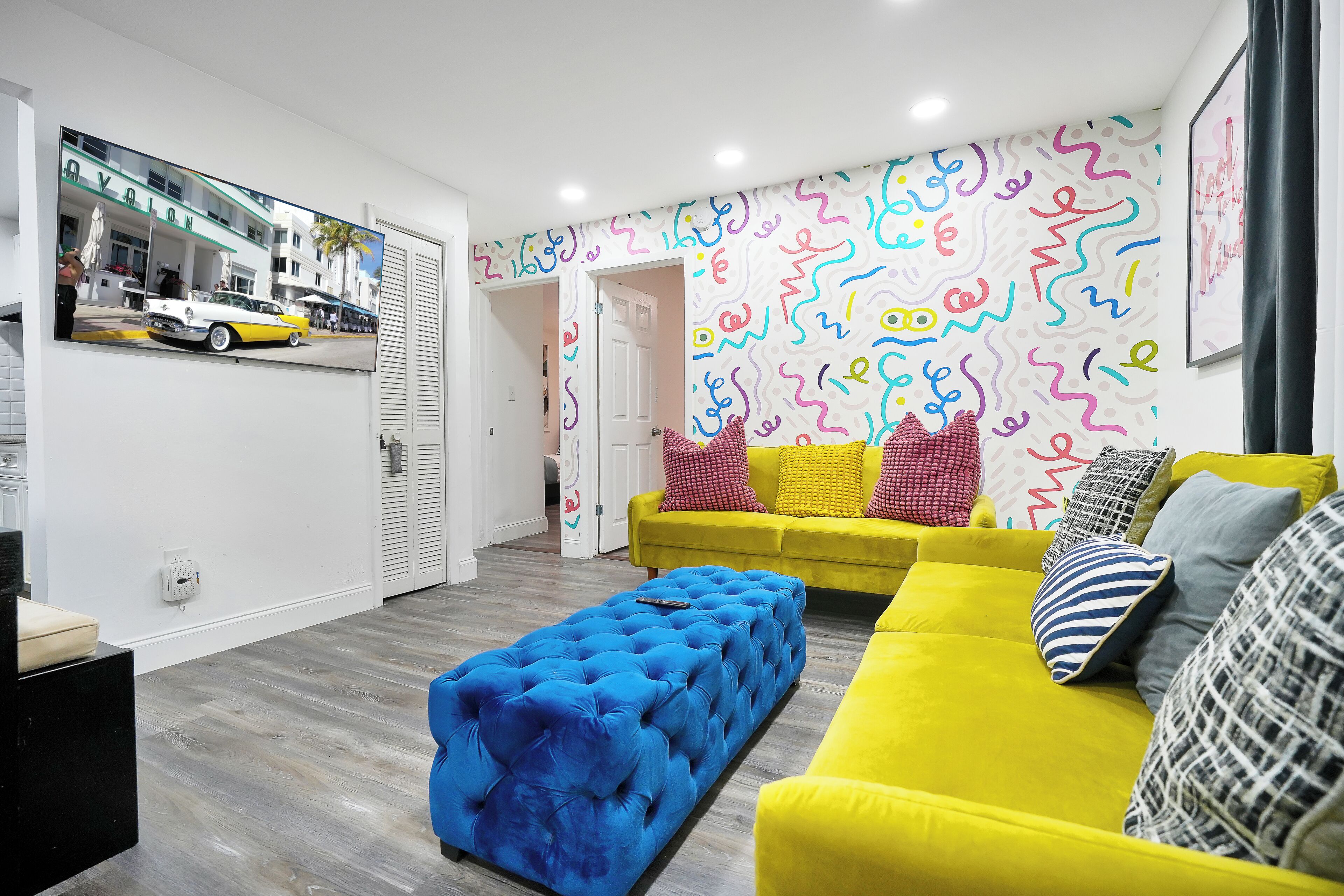 Una sala moderna con un sofá amarillo, un reposapiés azul y un llamativo mural abstracto.