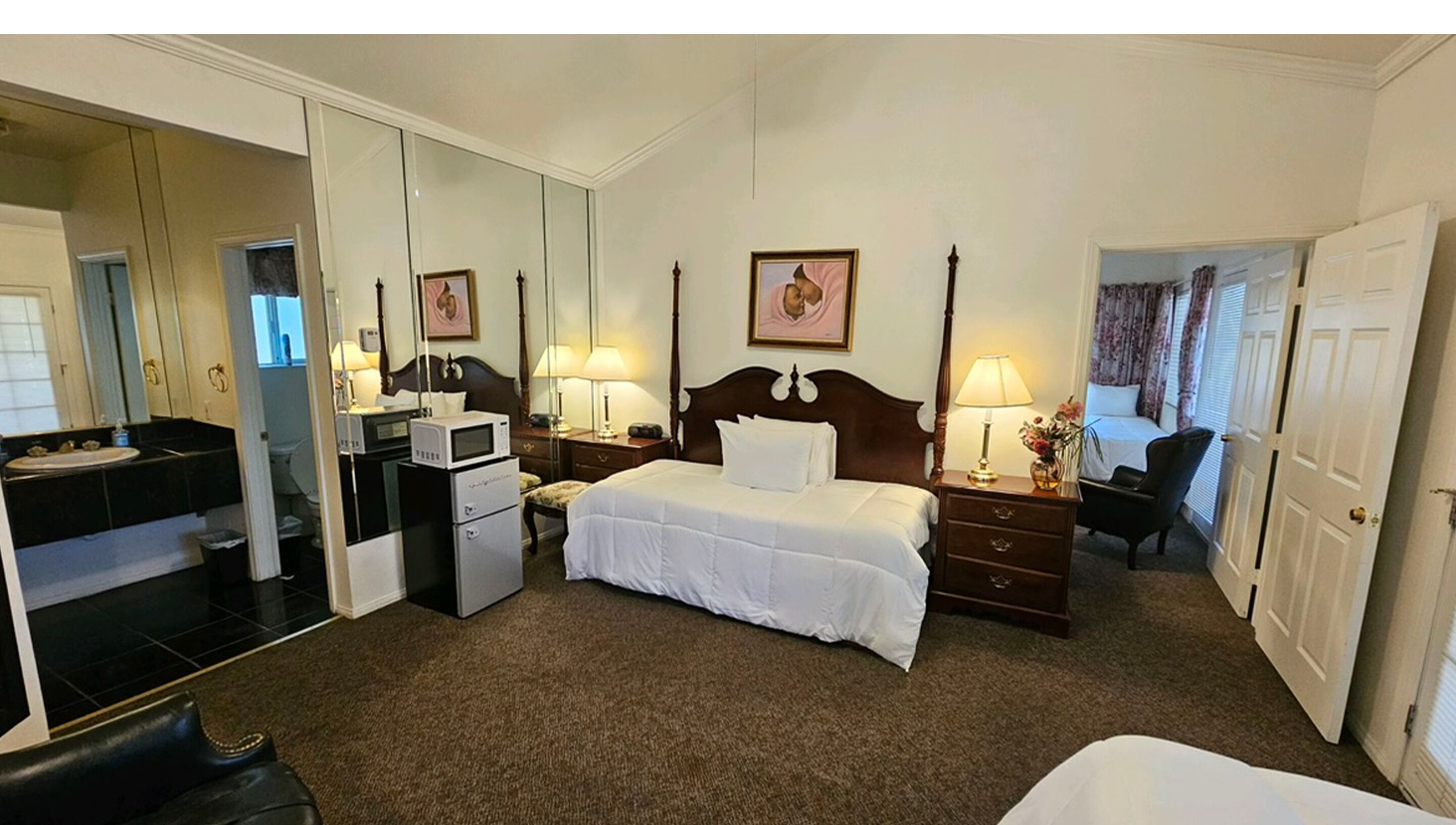 A hotel room with a large bed, a bedside table with a lamp, a small desk with a chair, a mirror, a wardrobe, and a bathroom with a sink and a toilet.