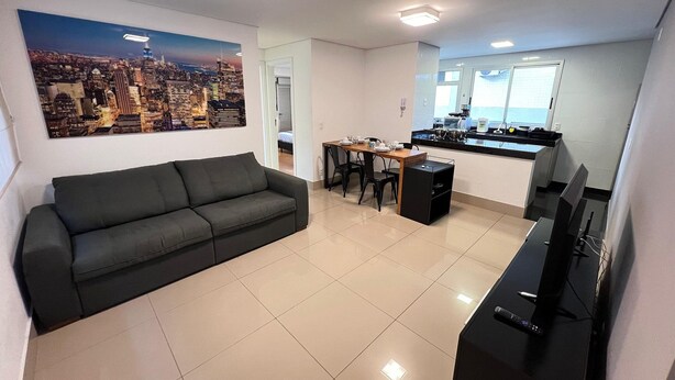 A modern living room with a black sofa, a dining table with chairs, a kitchen area with a TV, and a cityscape painting on the wall. - Lockings  Lourdes 12 (Belo Horizonte)