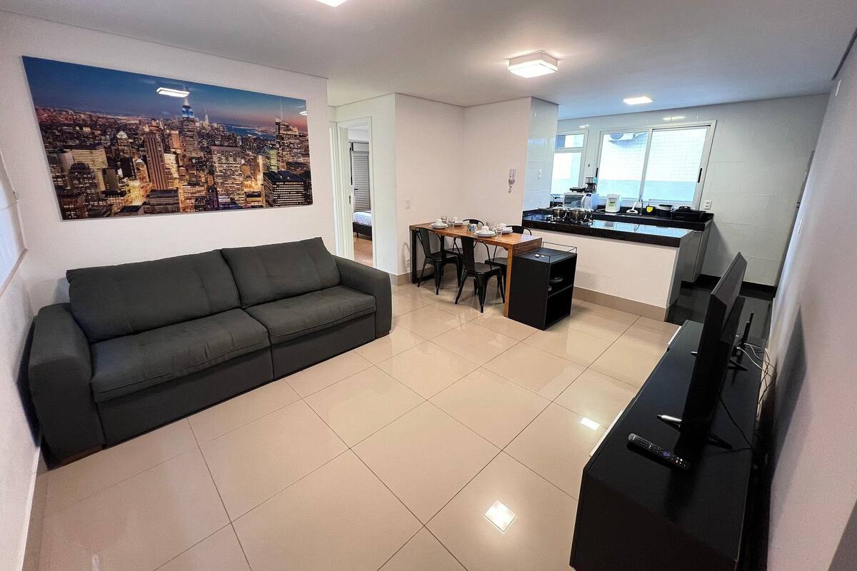 A modern living room with a black sofa, a dining table with chairs, a kitchen area with a TV, and a cityscape painting on the wall.