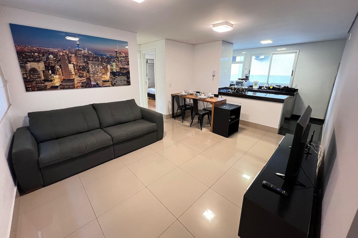 Una sala de estar moderna con un sofá negro, una mesa de comedor con sillas, un área de cocina con televisor y un cuadro de un paisaje urbano en la pared.