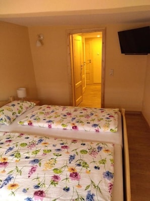 A bedroom with a bed covered in floral bedding, a wooden floor, a wall-mounted TV, and a door leading to a hallway.