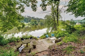 A lakeside patio with stone tables and chairs, surrounded by lush greenery and a view of a bridge. - Upper Black Eddy Home w/ Delaware River Access! (Upper Black Eddy)