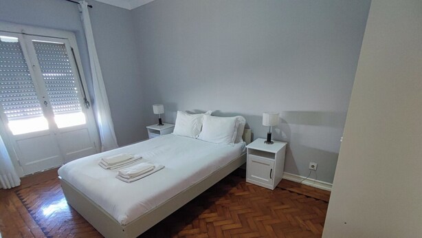 A bedroom with a bed, two bedside tables, a nightstand, a door, and a window with blinds. - Lisbon traditional T2 with balcony in Damaia (Amadora)