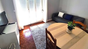 A living room with a sofa, a dining table with chairs, a TV on a stand, and a window with blinds. - Lisbon traditional T2 with balcony in Damaia (Amadora)