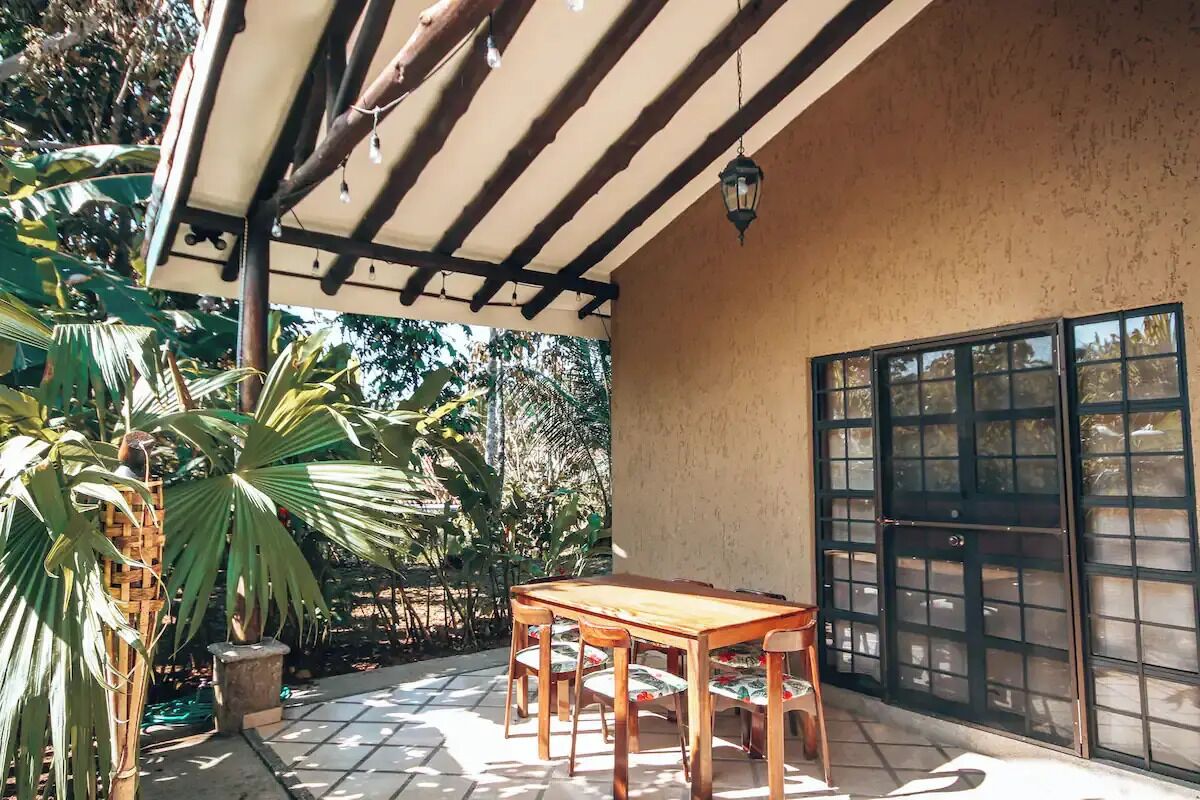 A covered patio with a wooden table and chairs, surrounded by lush greenery and a large window with a grid pattern.