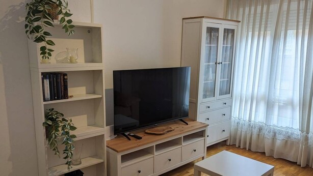 Un salon avec un téléviseur posé sur un meuble en bois, des étagères blanches, un buffet et une fenêtre ornée de rideaux légers.