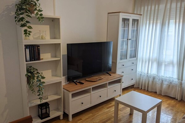 Un salon avec un téléviseur posé sur un meuble en bois, des étagères blanches, un buffet et une fenêtre ornée de rideaux légers.