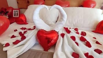Una cama con una toalla dispuesta en forma de corazón, pétalos de rosa roja y almohadas con forma de corazón.