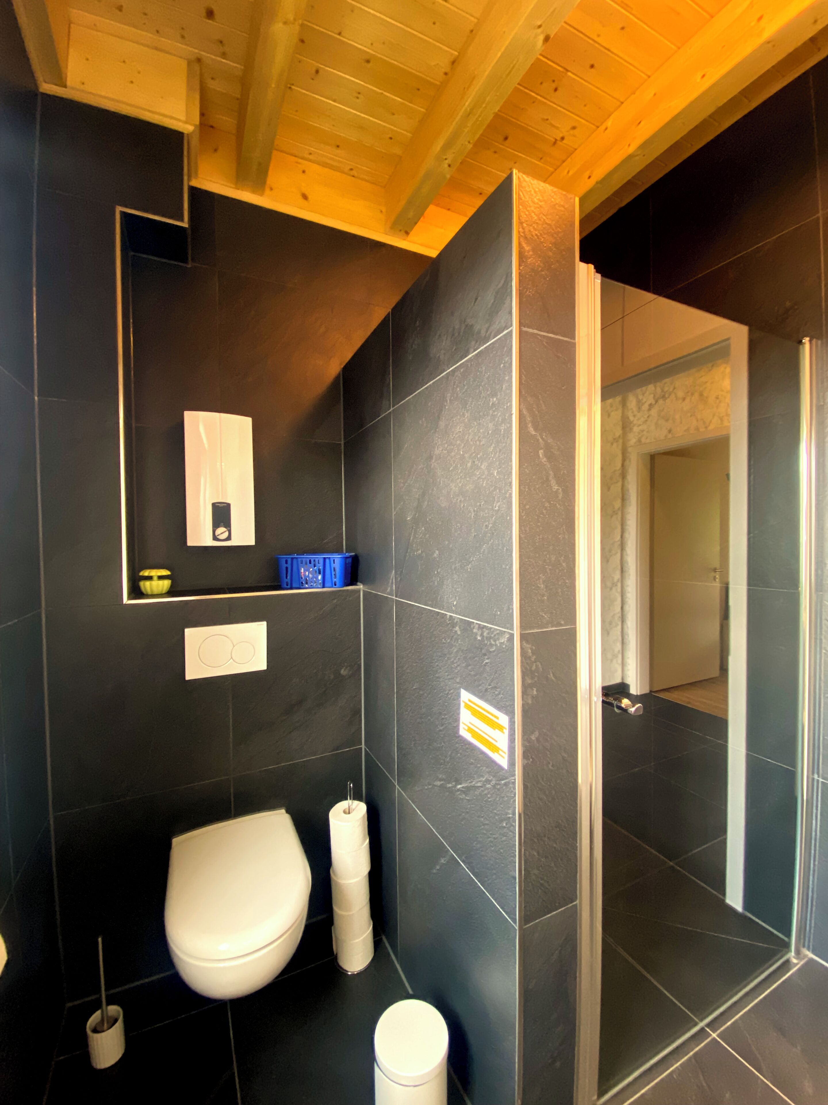 Une salle de bain moderne avec des toilettes blanches, des carreaux foncés, un espace douche et des poutres en bois au plafond.