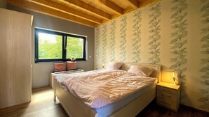 Een slaapkamer met een houten plafond, een bed met witte beddengoed, een nachtkastje met een lamp en een raam met uitzicht op bomen.