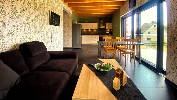 Een moderne woonkamer met een donkerbruine bank, een houten salontafel en een eethoek met houten stoelen en een tafel. Achtergrond is een keuken met witte kasten en een roestvrijstalen koelkast.