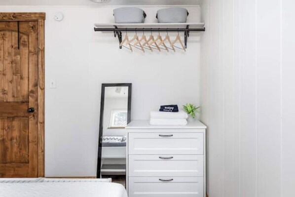 A bedroom with a wooden door, a white dresser with drawers, a mirror, and a shelf with cushions and hangers.