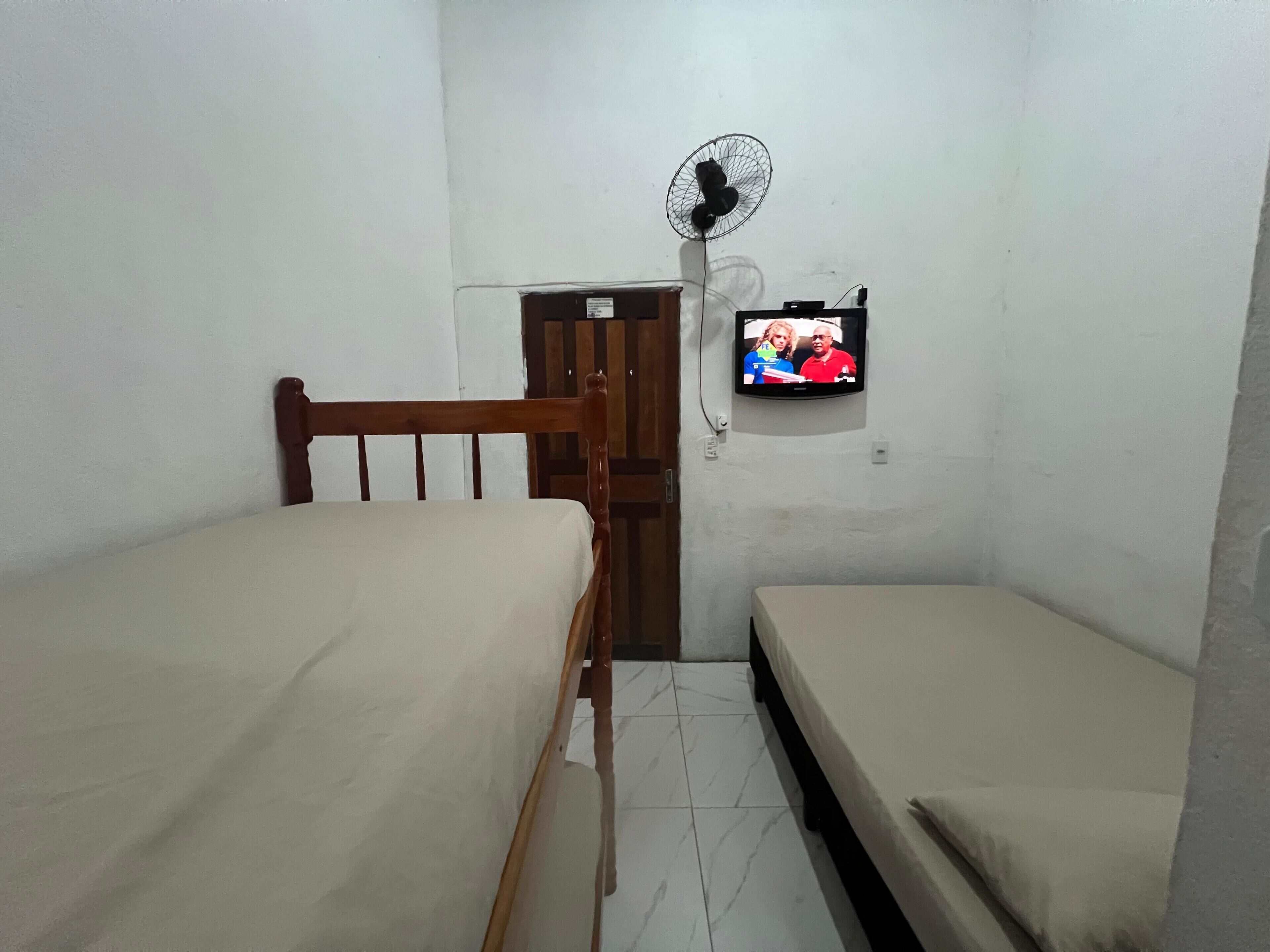 A small, dimly lit room with two wooden beds, a single wall-mounted TV, and a standing fan.