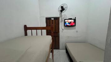 A small, dimly lit room with two wooden beds, a single wall-mounted TV, and a standing fan.