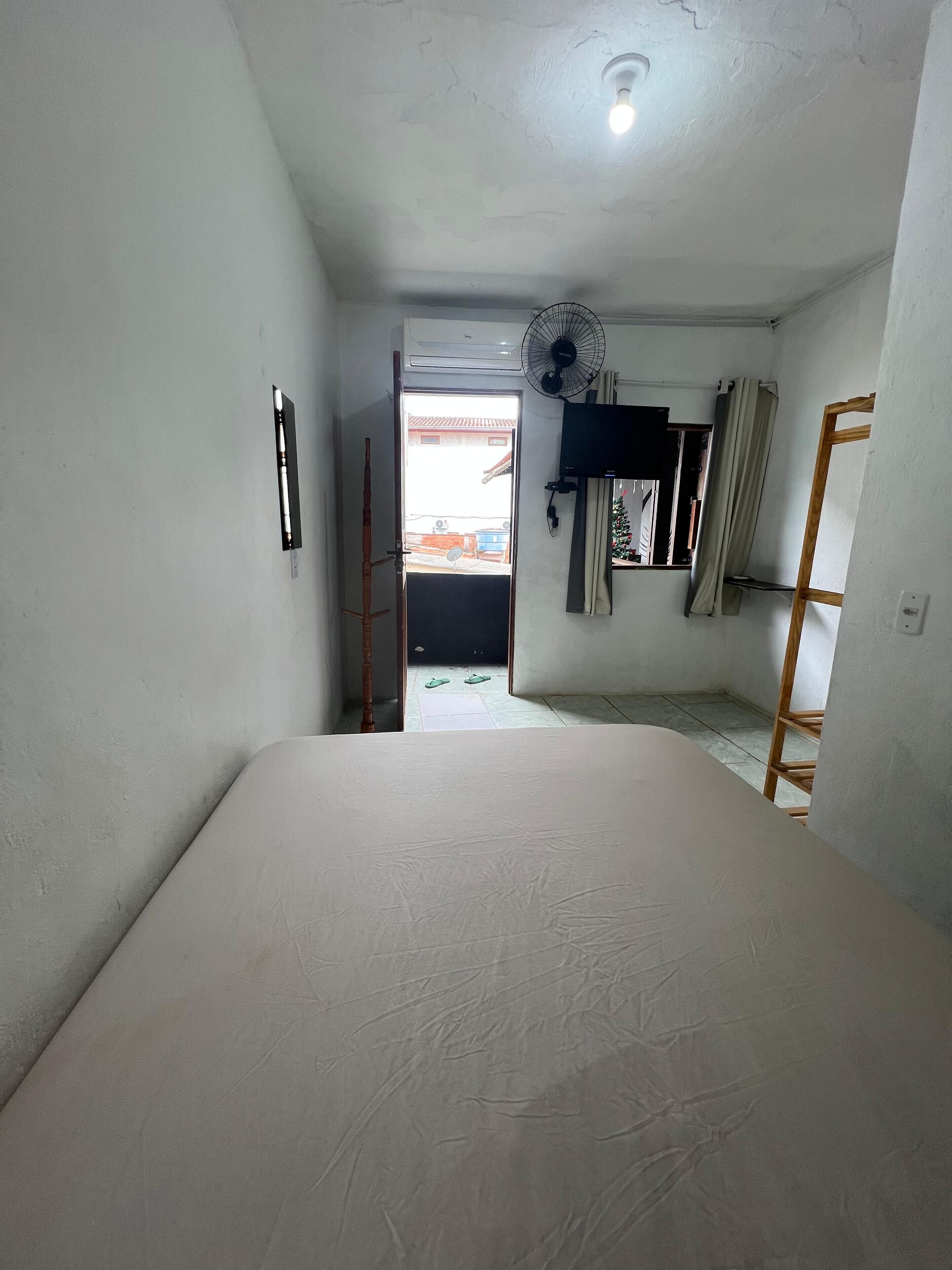 A narrow room with a bed, a fan, a TV, and a window with curtains.