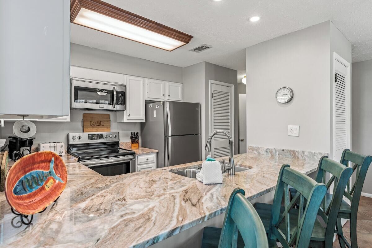 A modern kitchen with stainless steel appliances, a marble countertop, and teal chairs.