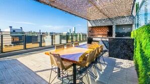 En udendørs spiseplads med et stort bord og stole, grill og udsigt til byens skyline.