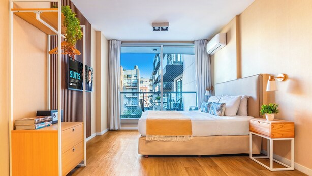 Et moderne hotelværelse med en stor seng, en trækommode, et lille sengebord og udsigt over byen gennem et stort vindue.