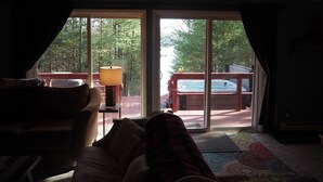A cozy room with a wooden ceiling, a large window, and a view of a hot tub outside. - The Cub Hill Chalet on Cub Lake - Private Lakefront, Dock, Raft, Water Toys, Huge Deck, and Spa! (Kalkaska)