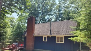 A small, single-story building with a dark blue exterior, a brick chimney, and a sloped roof. - The Cub Hill Chalet on Cub Lake - Private Lakefront, Dock, Raft, Water Toys, Huge Deck, and Spa! (Kalkaska)