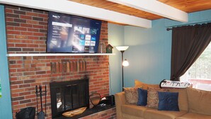A living room with a brick fireplace, a flat-screen TV mounted above it, a sofa with patterned cushions, and a coffee table with a newspaper and remote control. - The Cub Hill Chalet on Cub Lake - Private Lakefront, Dock, Raft, Water Toys, Huge Deck, and Spa! (Kalkaska)