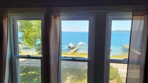 A room with three windows overlooking a body of water, a dock with chairs, and a grassy area.