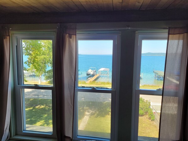 A room with three windows overlooking a body of water, a dock with chairs, and a grassy area.