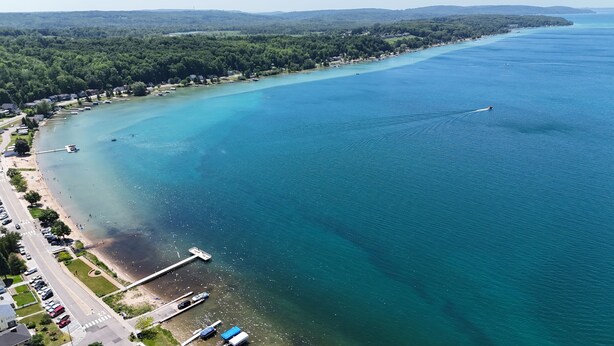 An aerial view of a coastal area with a marina, a road along the shore, and a forested landscape in the background.