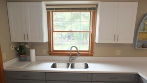 A kitchen with a double sink, grey cabinets, a window above the sink, and a potted plant on the counter. - The Maple Cottage - WALK TO BEACH, LAKE MICHIGAN, AND DOWNTOWN! (Frankfort)
