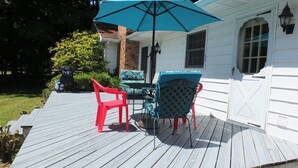 A wooden deck with a table and chairs, a blue umbrella, and a white building with a window and door. - The Maple Cottage - WALK TO BEACH, LAKE MICHIGAN, AND DOWNTOWN! (Frankfort)