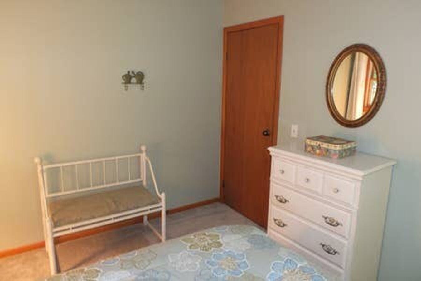 Un dormitorio con una cama de metal blanco, una puerta de madera, un aparador con cajones y un espejo en la pared.