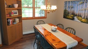 A dining area with a wooden table, chairs, a chandelier, a bookshelf, and a painting on the wall. - The Maple Cottage - WALK TO BEACH, LAKE MICHIGAN, AND DOWNTOWN! (Frankfort)