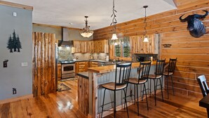 A rustic kitchen with wooden walls, a central island with bar stools, and a bull head mounted on the wall. - Weatherwood - Gorgeous Luxury Log Cabin with Spa, Sauna, & Games!  8 minutes from Crystal Mountain! (Copemish)