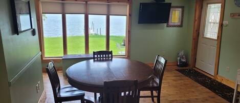A dining area with a round table and four chairs, large windows with a view of a grassy area and water, a flat-screen TV mounted on the wall, and a wooden door with a glass panel.