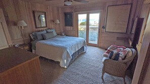 A wooden cabin bedroom with a bed, a chair, a desk, and a sliding glass door leading to a balcony.