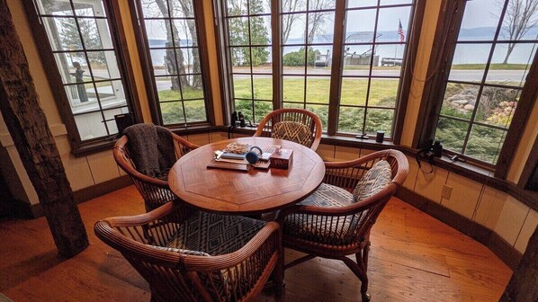 A corner room with a large window overlooking a grassy area and a body of water. There is a round wooden table with chairs and a few items on it.