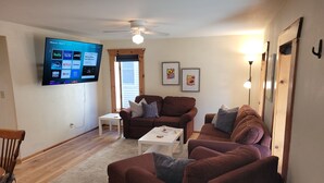 A living room with a flat-screen TV mounted on the wall, a ceiling fan, and a window with blinds. There are two couches, a coffee table, and a wooden chair. - The Coral Castle - Large, Newly-Remodeled, Gorgeous, and Great Location!  Walk to Beach and Downtown! (Frankfort)