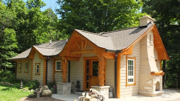 Une cabane en rondins à deux étages, dotée d’une cheminée en pierre, d’un porche en bois et d’une allée gravillonnée.