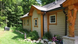 Une maison en bois à deux étages, dotée de fondations en pierre, entourée d’un jardin bien entretenu où poussent diverses plantes et fleurs.