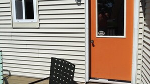 Een klein huisje met één deur en één raam, een veranda met een stoel, en een lantaarnpaal.