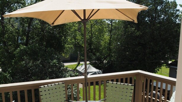 Een ronde glazen tafel met een beige parasol, omringd door vier rieten stoelen op een houten terras met een balustrade.