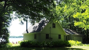 A green house with a sloped roof, surrounded by lush greenery and a lake view. - A Short's Walk - Sandy Lakefront with Spa / Hot Tub and Kayaks! (Elk Rapids)