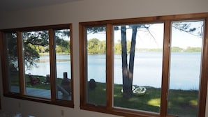 A kitchen with large windows overlooking a lake, a wooden deck, and outdoor seating. - A Short's Walk - Sandy Lakefront with Spa / Hot Tub and Kayaks! (Elk Rapids)