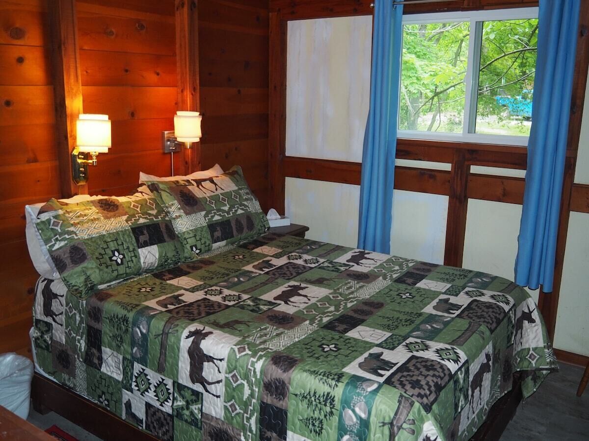 Un dormitorio de cabaña de madera con una cama cubierta por un edredón estampado, dos lámparas fijadas a la pared y una ventana con cortinas.