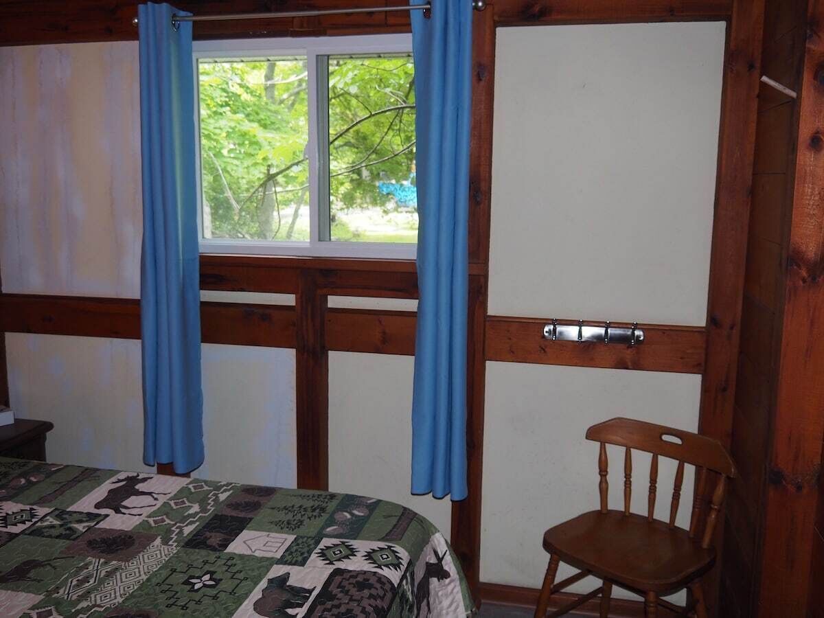 A room with a bed, a wooden chair, a window with blue curtains, and a door.