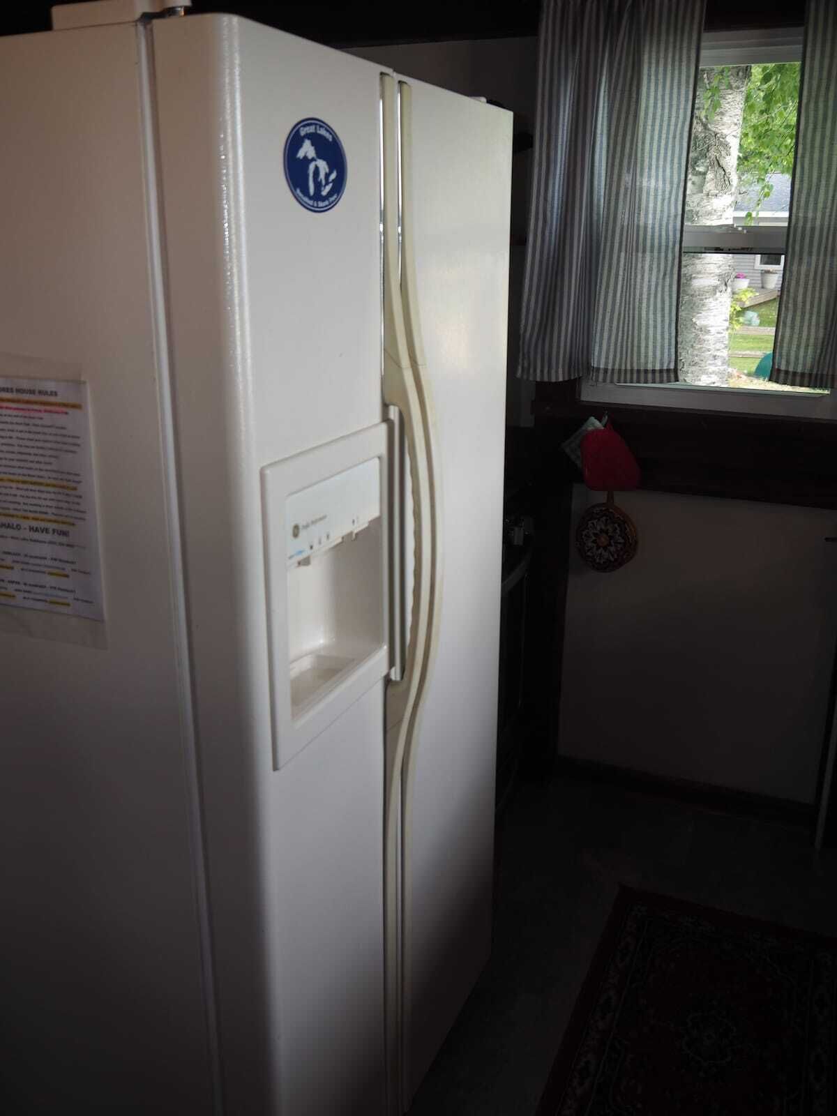 A compact kitchen with a white refrigerator, a small window with curtains, and a visible outdoor view.